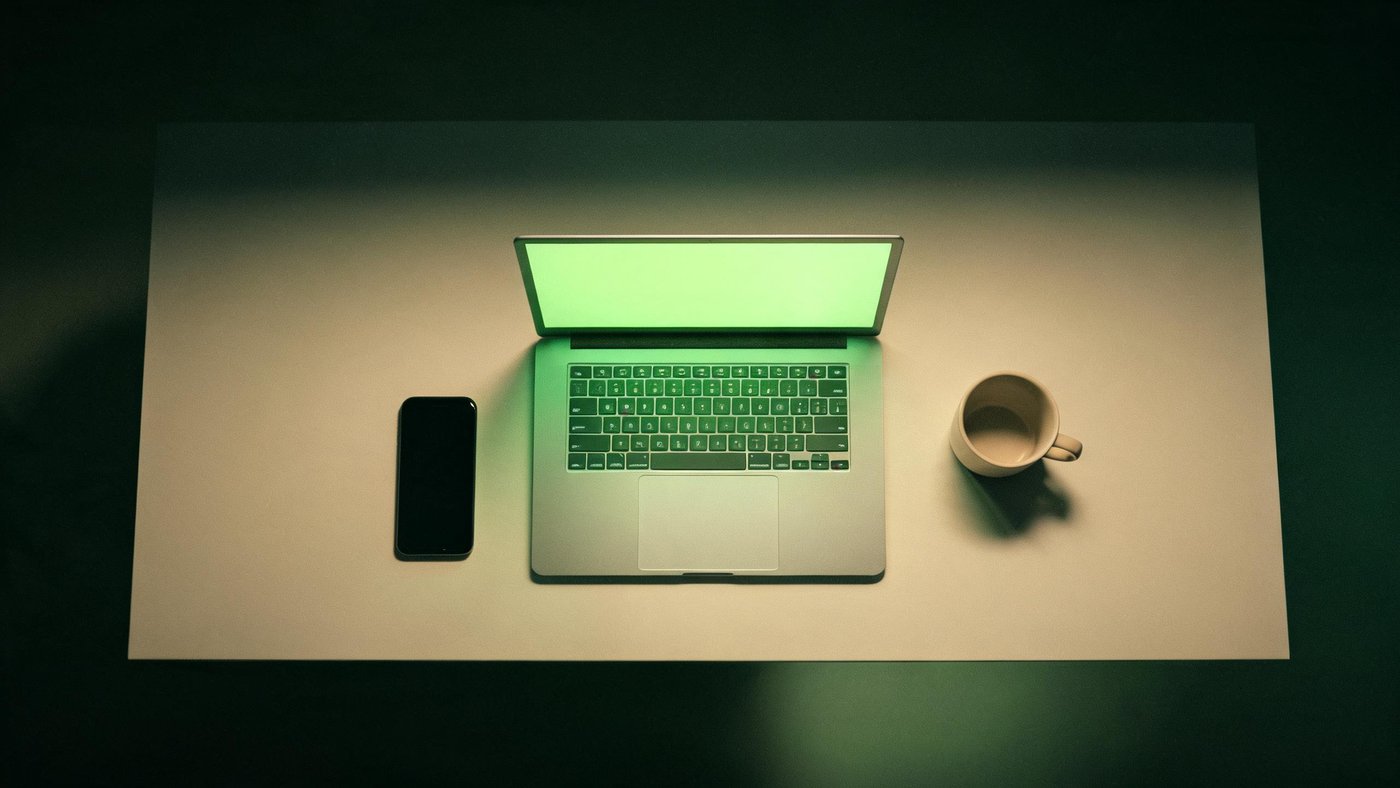 A dark desk at night lit by an open laptop with a soft green glow, an empty coffee cup beside it